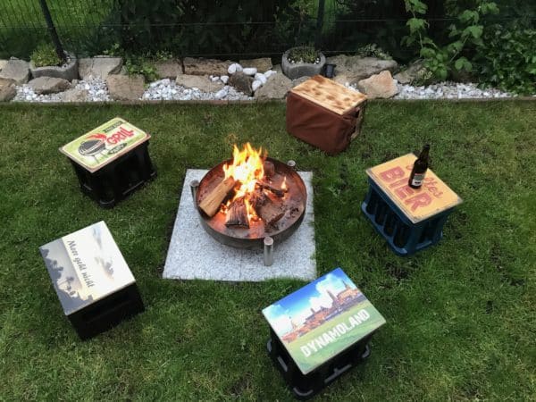 Eine Feuerstelle in einem Garten mit diversen Bierkastensitzen die in einem Kreis um das Lagerfeuer stehen. Auf dem Motiv kaltes Bier ist eine vermeintliche Bierflasche platziert.