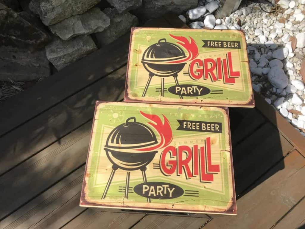 zwei Bierkastensitze mit dem Druck GrillParty mit schwarzen Bierkästen. Die Bretter liegen versetzt paralell zueinander und stehen im Garten. Sie werden in einen zarten goldenen Schimmer des Sonnenscheins gehaucht. frontalansicht nahaufnahme.